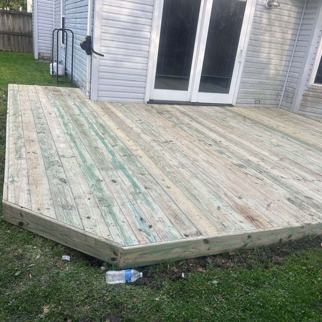 Wooden deck with a beveled edge, next to a house with glass doors, on a grassy lawn.