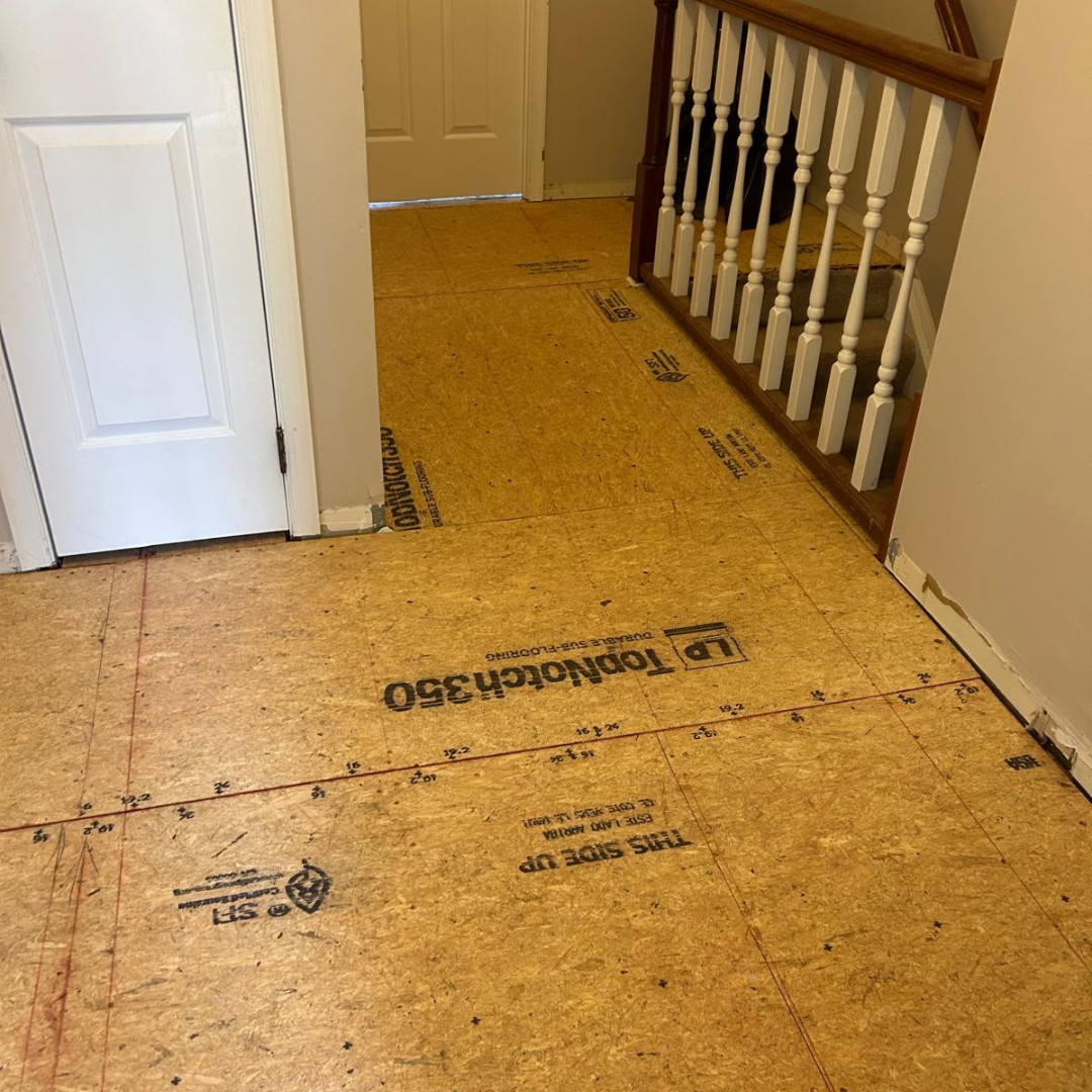 Hallway with new subfloor installed. White door and staircase with wood banister on the right.