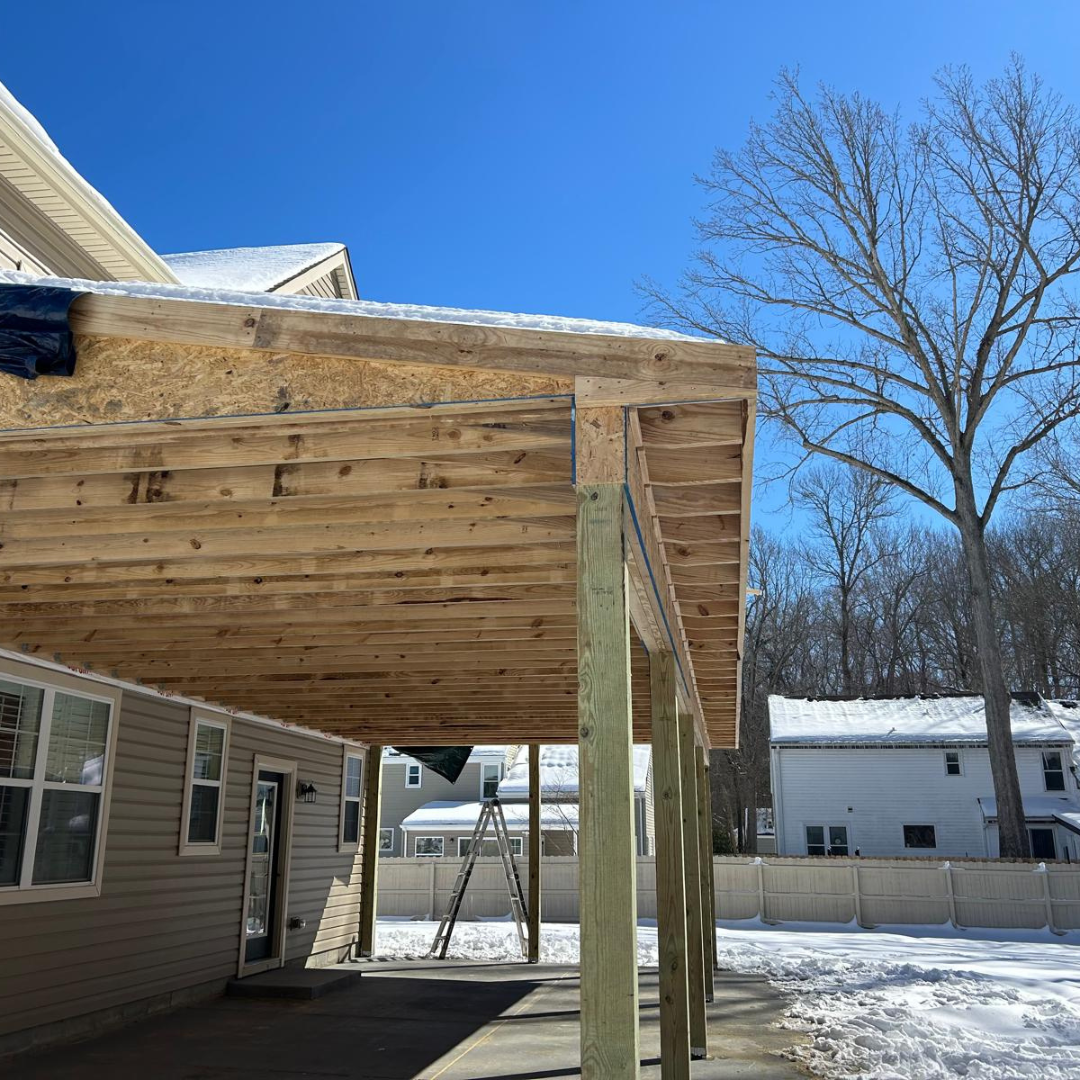 Partially built covered patio with exposed wood, a light-colored house, and a snowy backyard.