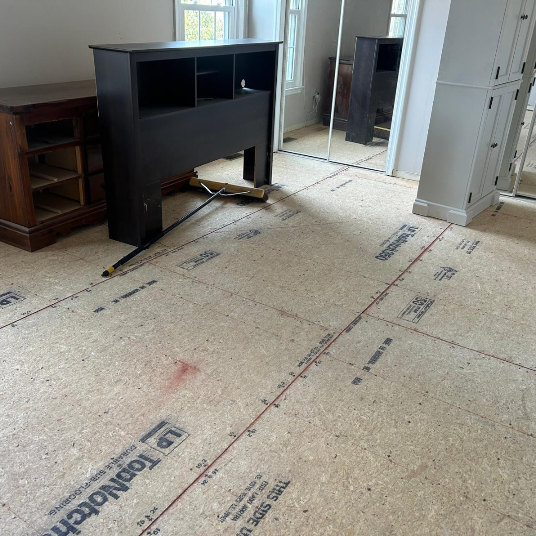 Room with new flooring in progress; a dark headboard and cabinet sit on top of the underlayment.