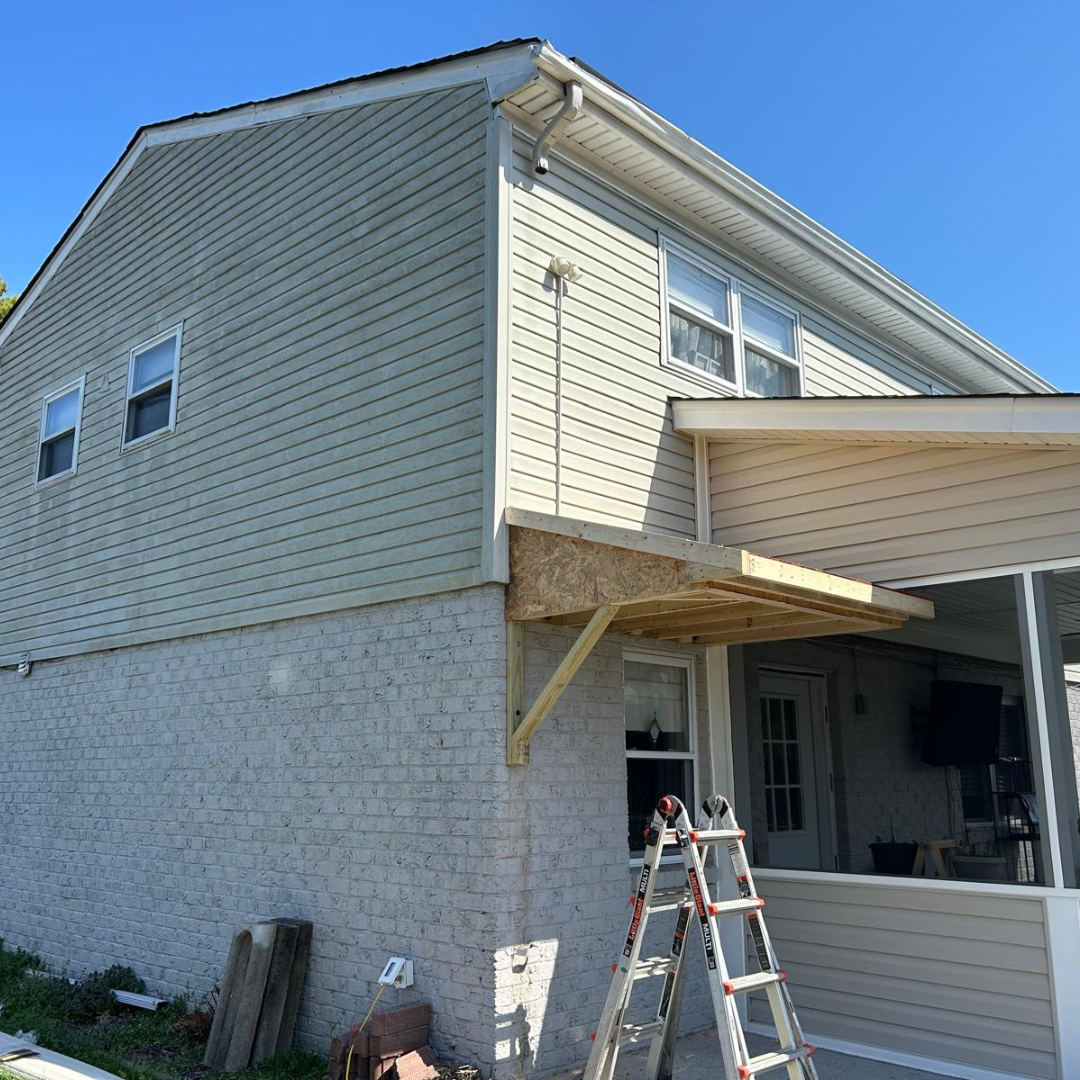 Two-story house under construction: gray siding, brick, scaffolding, and new wood awning over the porch.