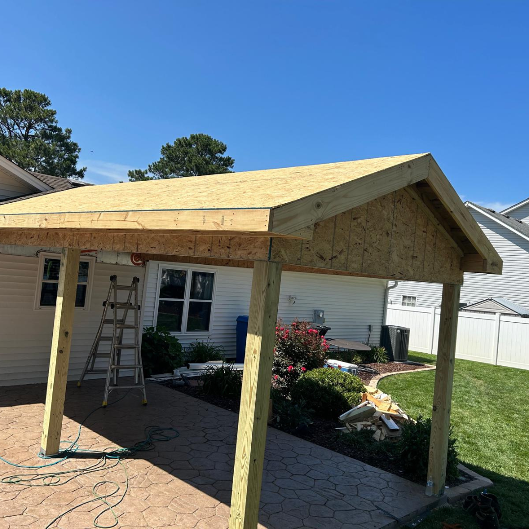 Carport under construction attached to a white house, featuring a ladder and blue sky.