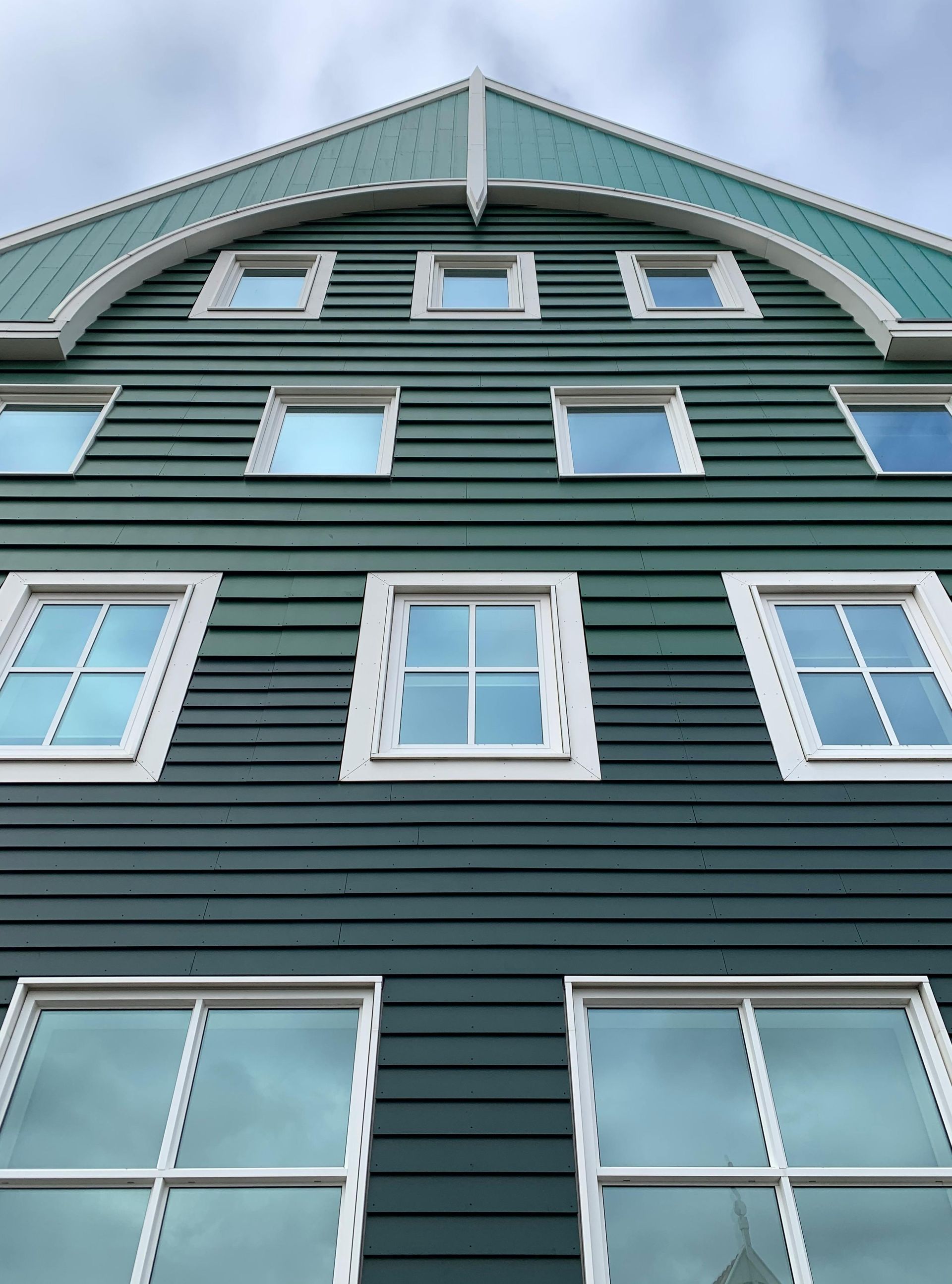 Green wooden building with white-framed windows under an arched roof, against a cloudy sky.