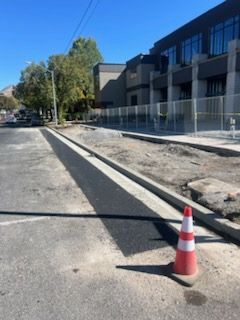 Road construction: Fresh asphalt strip with traffic cone in foreground, building and fence in background.
