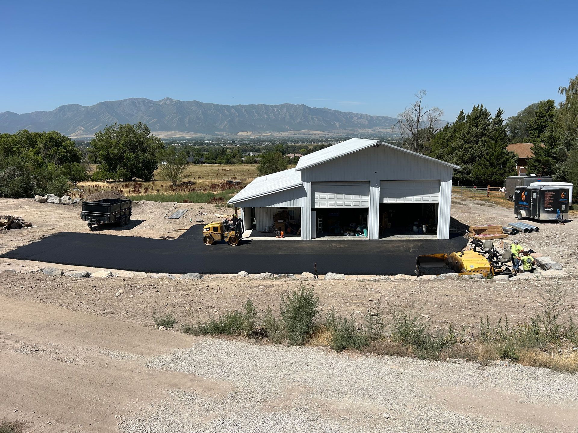 A paved driveway leads to a gray garage with open bays. Mountains and blue sky are in the background.