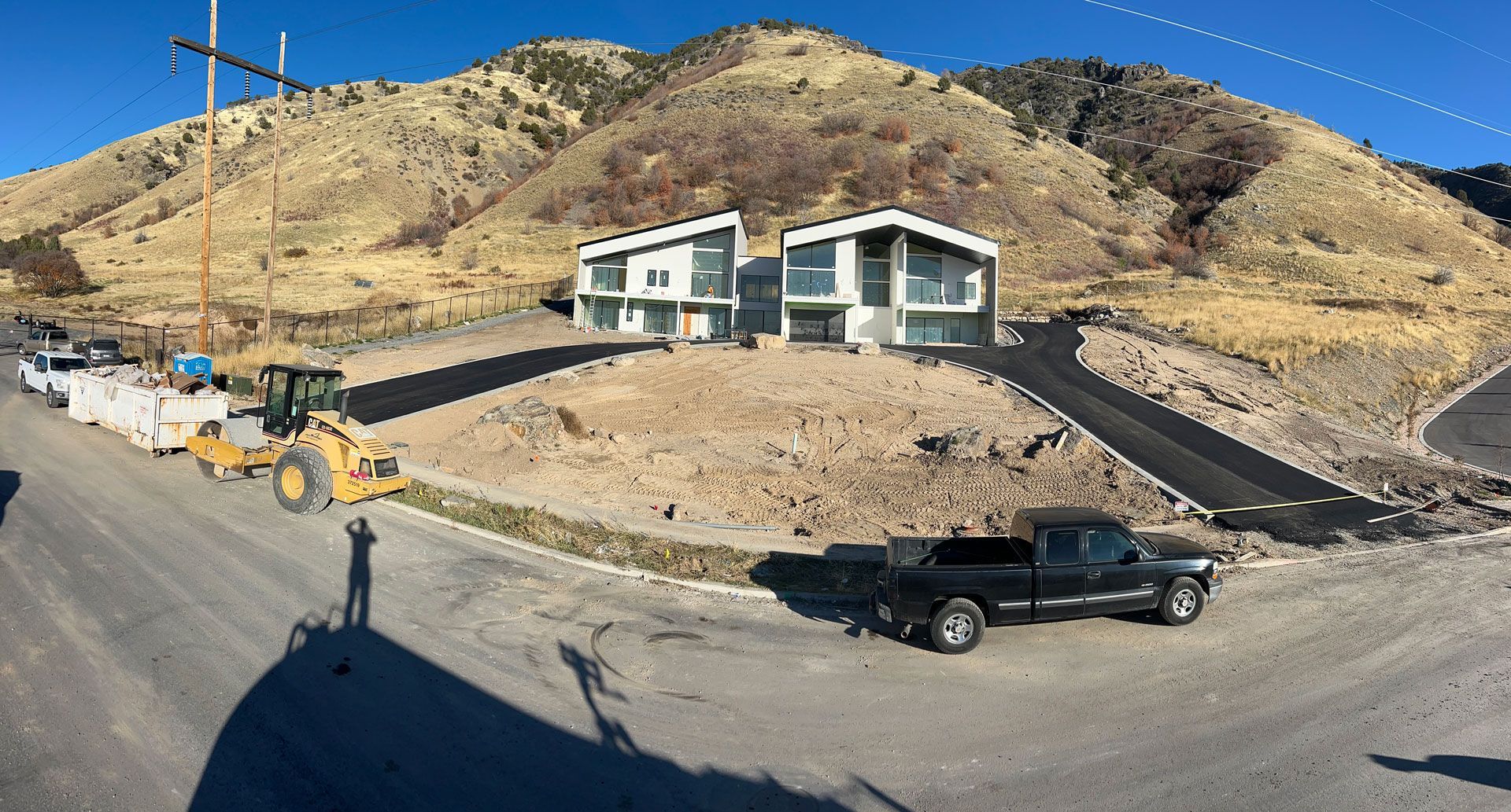 Modern homes on a hillside with construction equipment and a pickup truck. Paved road and a mountain backdrop.