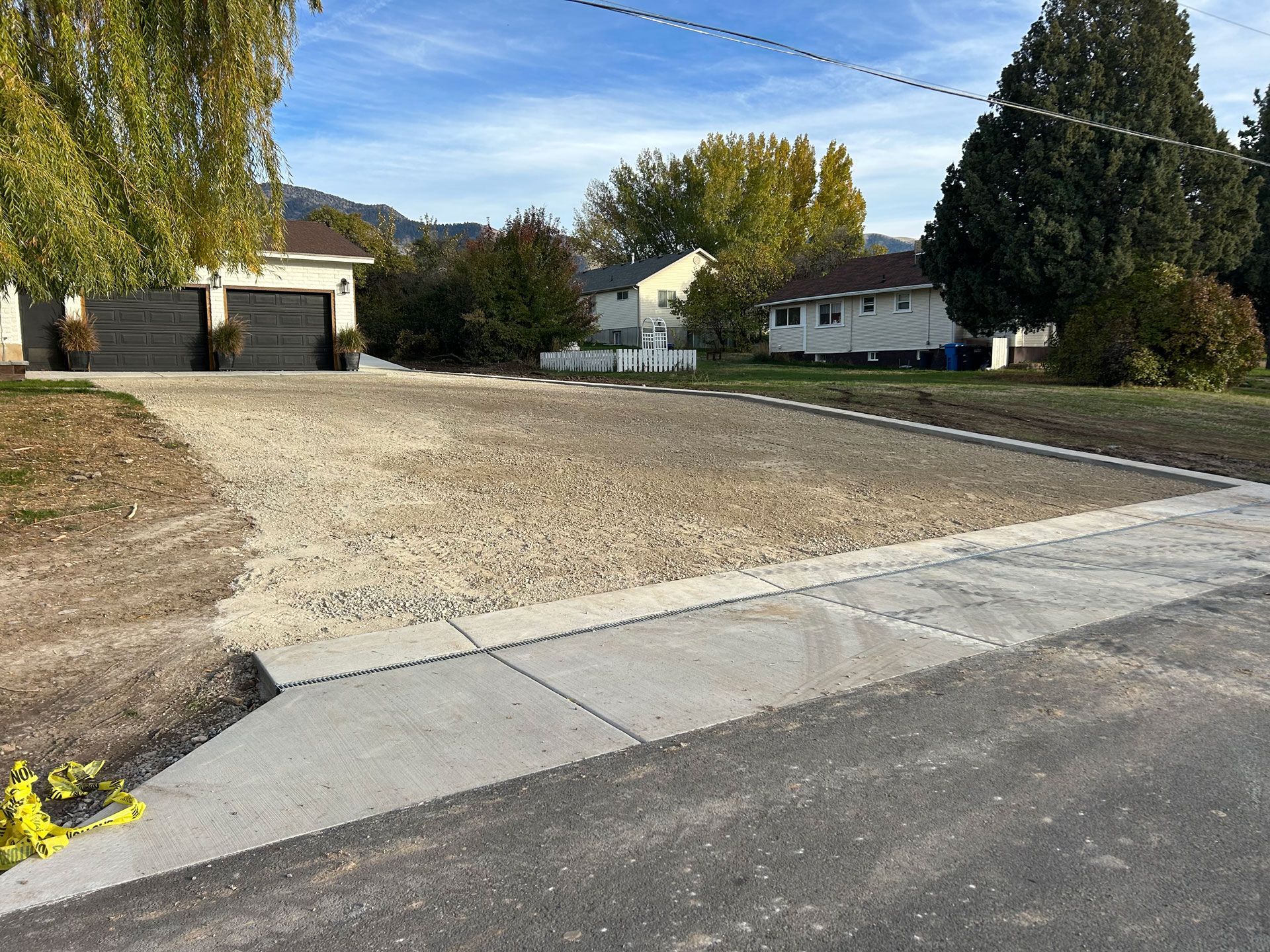 Gravel lot with concrete border, driveway, and detached garage. Houses and trees in the background under a blue sky.