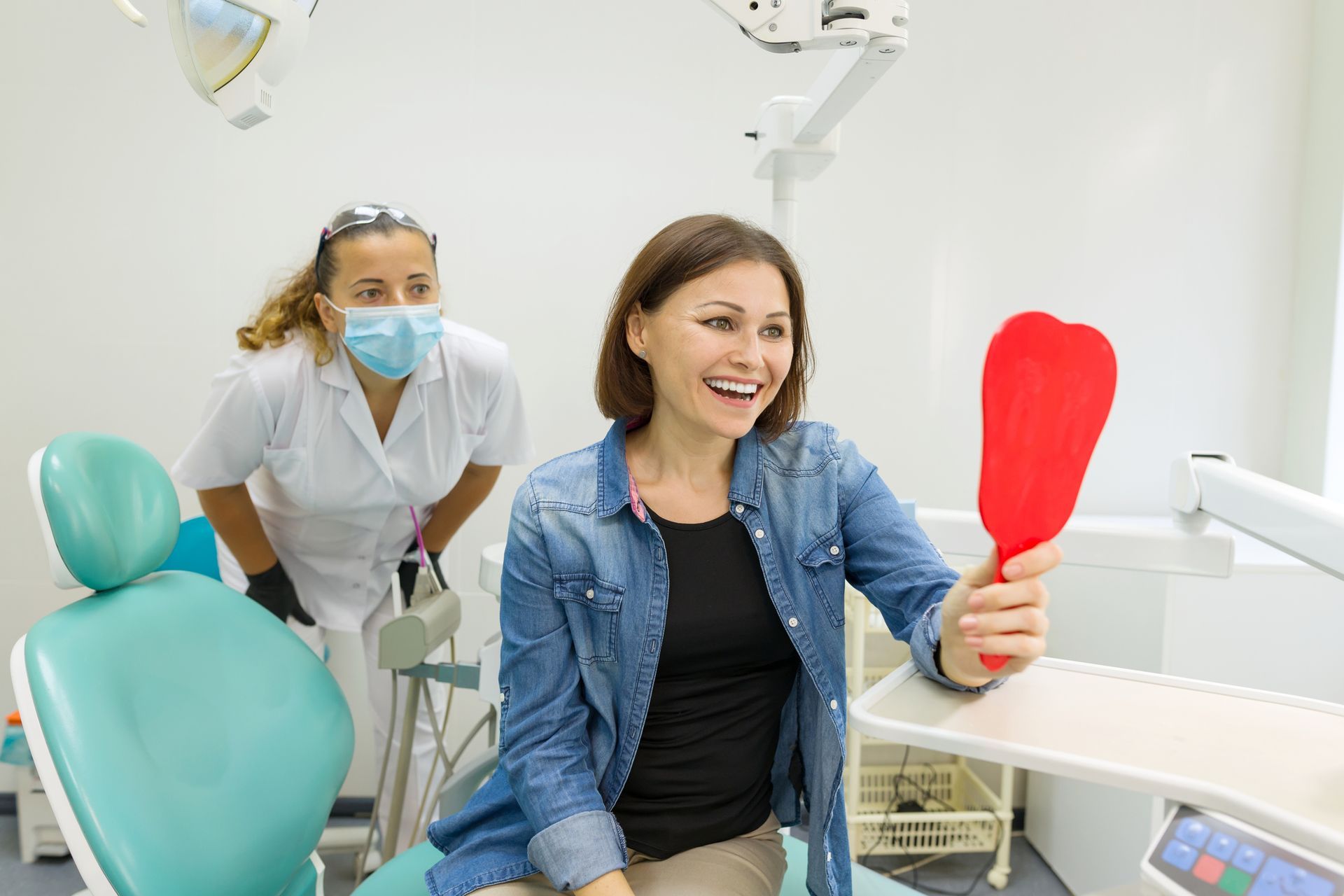 A woman is sitting in a dental chair looking at her teeth in a mirror.
