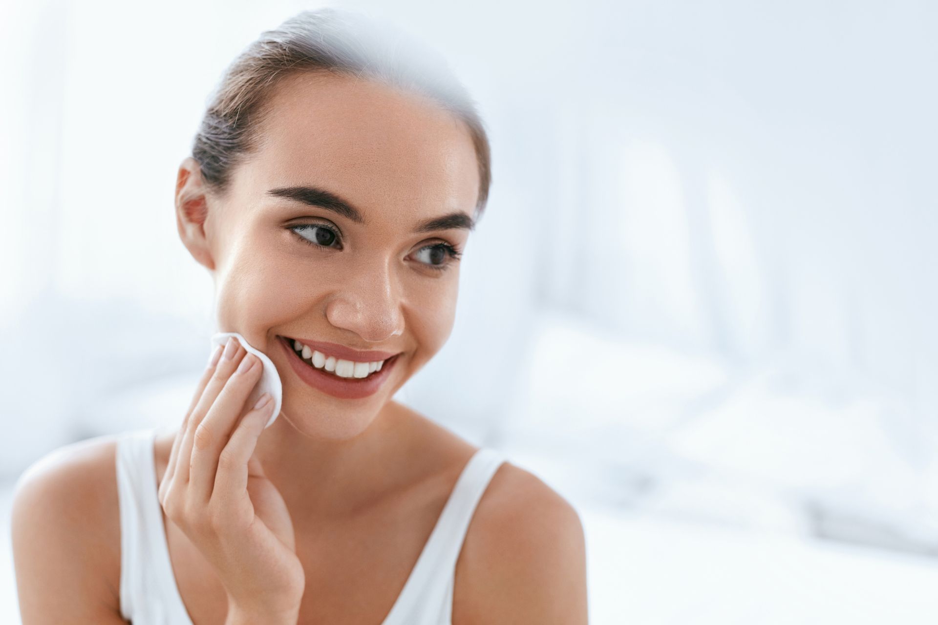 A woman is smiling while cleaning her face with a cotton pad.