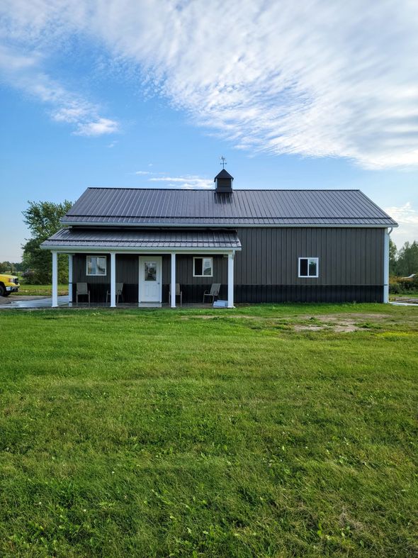 Gray barn-style building with white porch and black roof on a grassy field under a blue sky.
