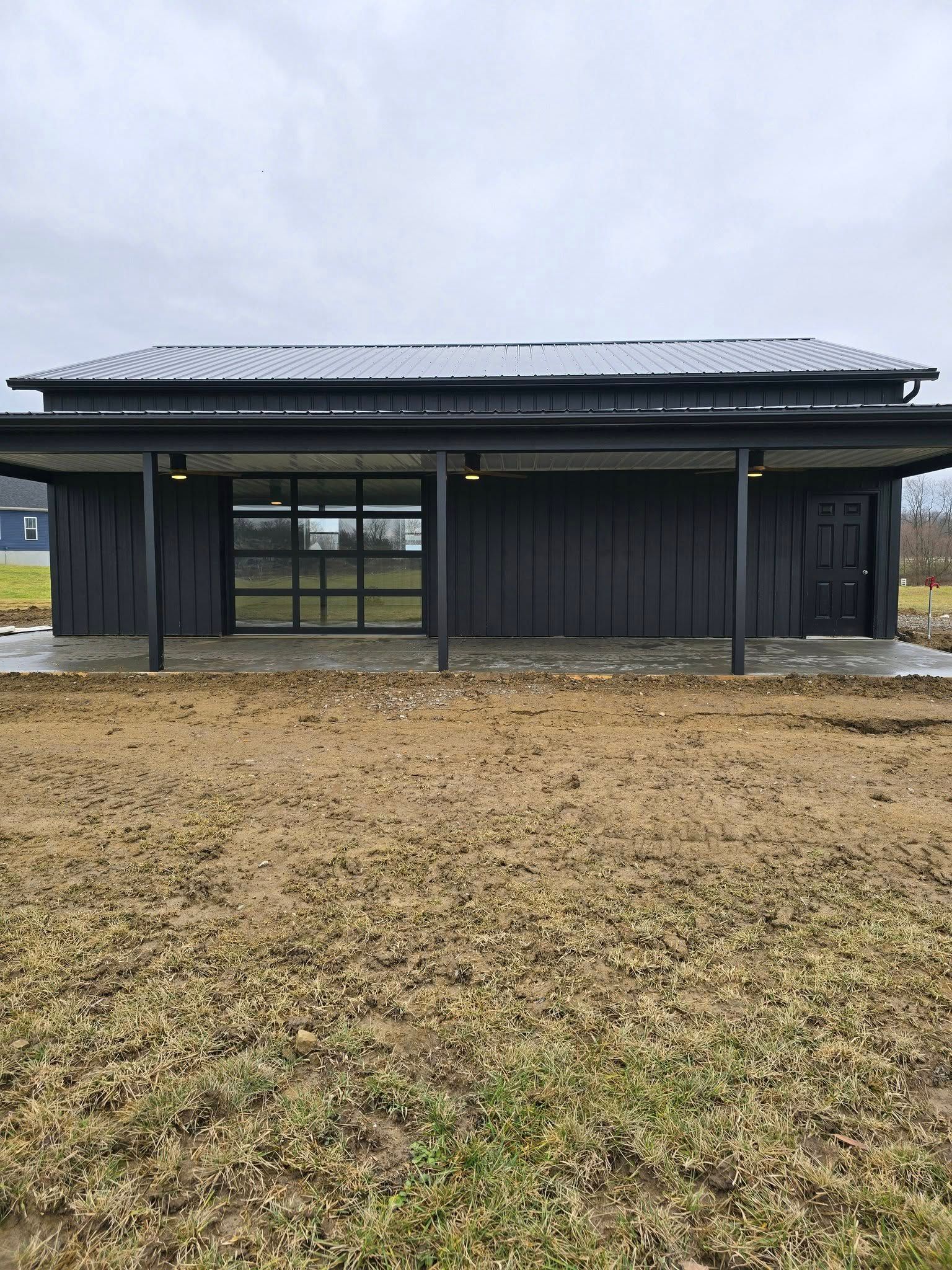 Black modern building with glass doors, porch, and a metal roof on a brown, muddy field.