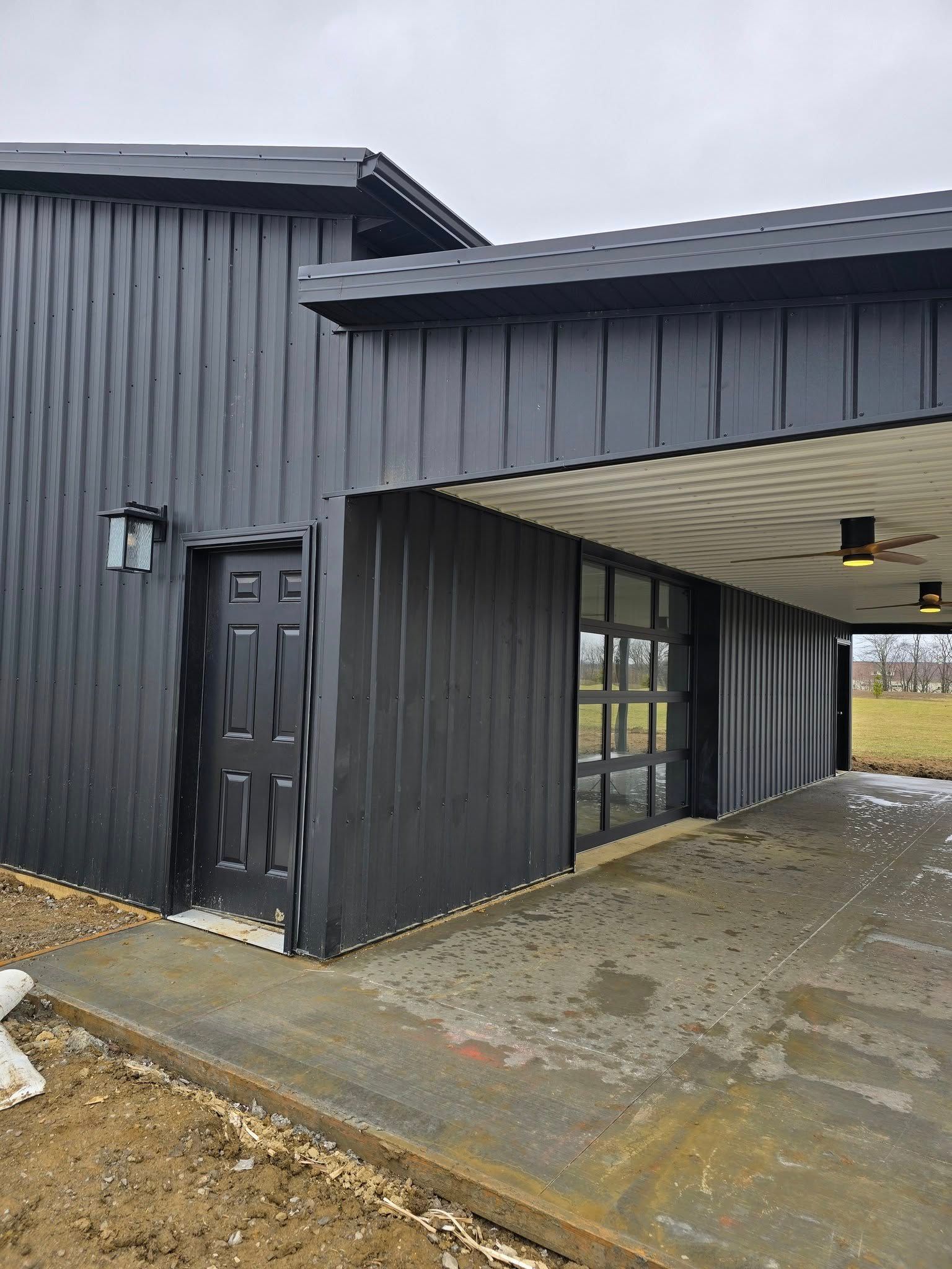 Black modern building with carport, door, and glass garage door. Concrete path.