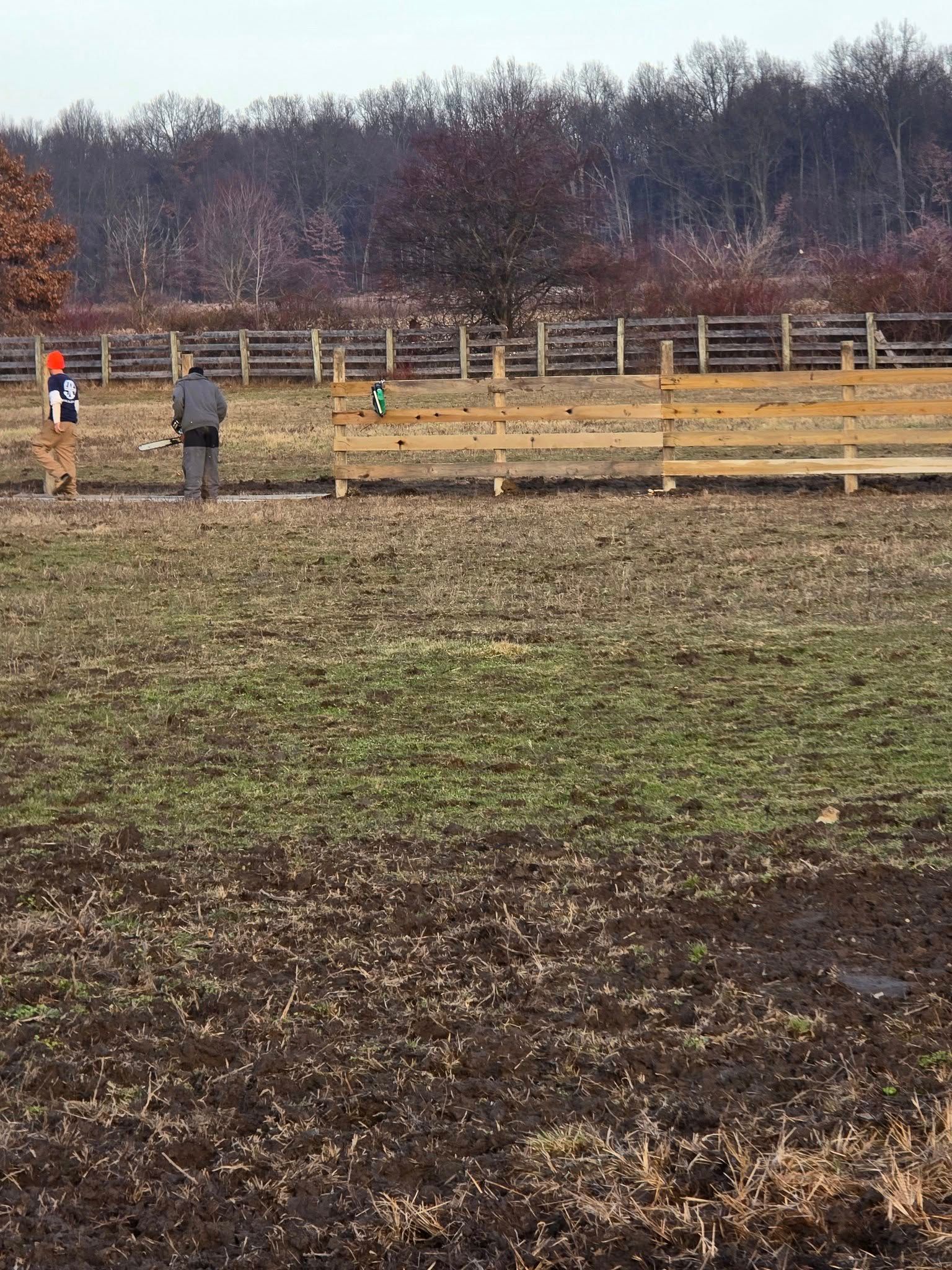 Two people in a field with a wooden fence, possibly target shooting. Bare trees in the background.
