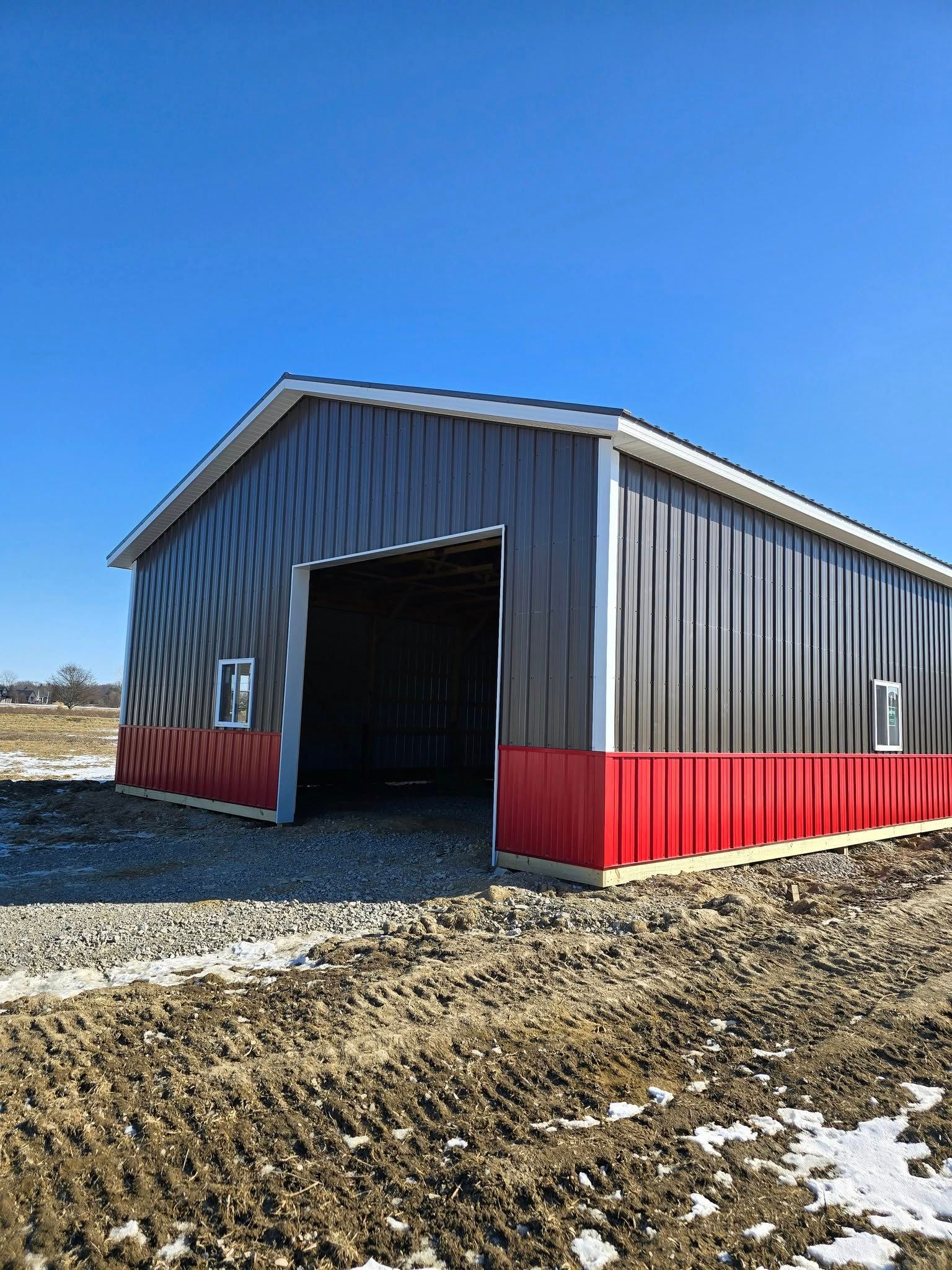 Gray and red metal barn on a sunny day with open door, in a muddy field.