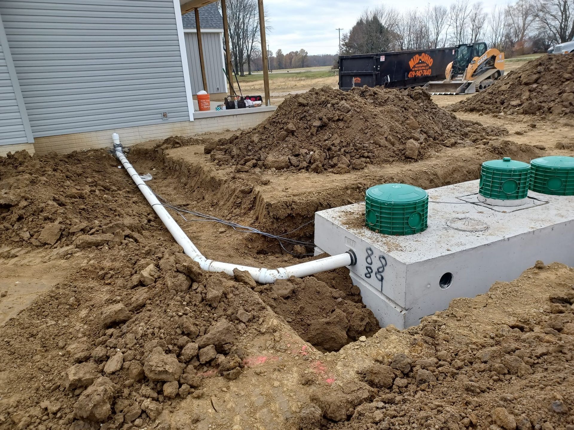 Installation of a septic system outside a light gray house with white trim.  Pipes are connected to a concrete tank.