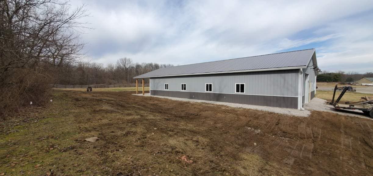 A light-colored metal building with a gray roof and a dirt field in front of it.