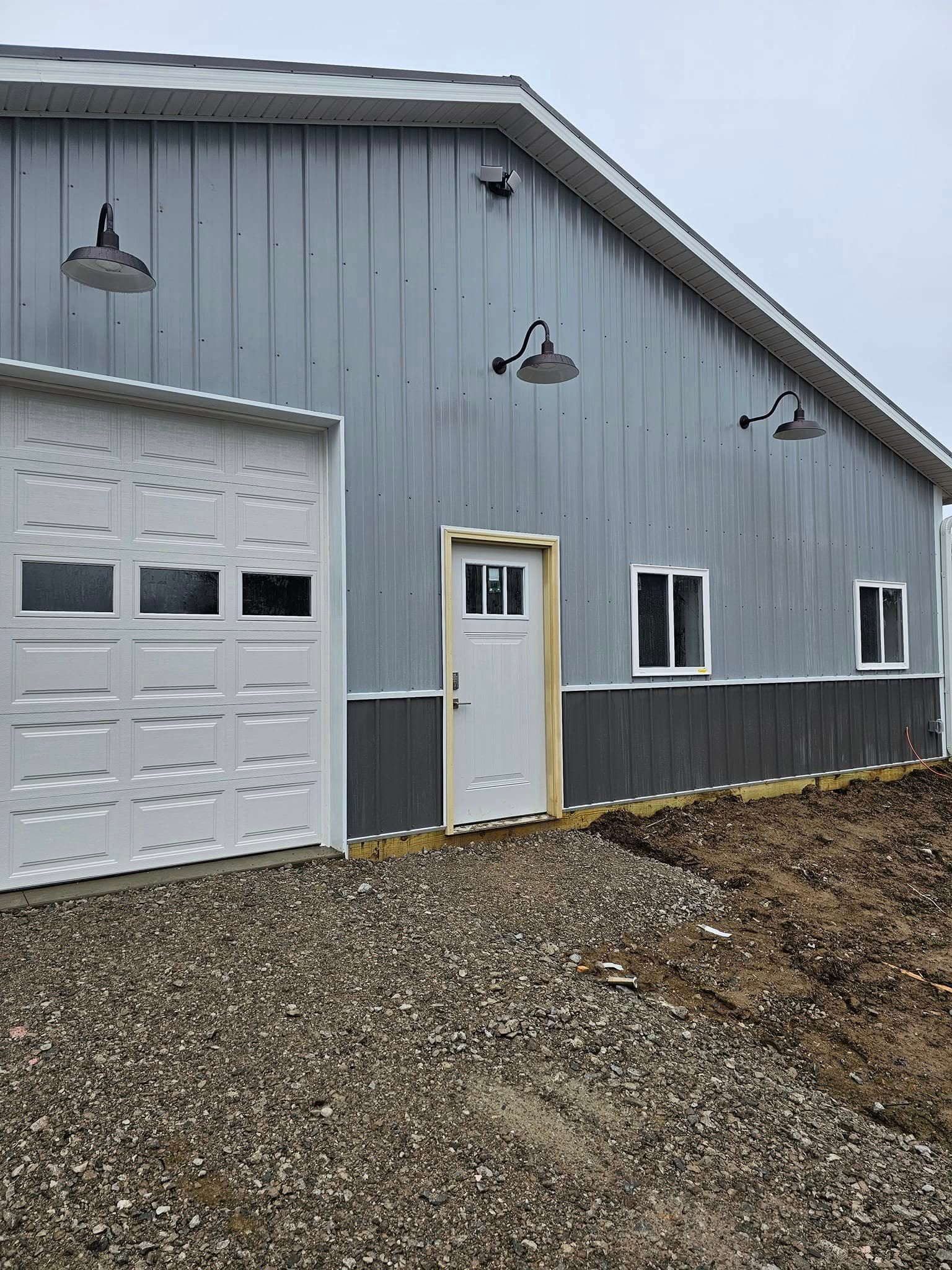 Gray metal building with garage door, white door, windows, and outdoor lights. Gravel ground.