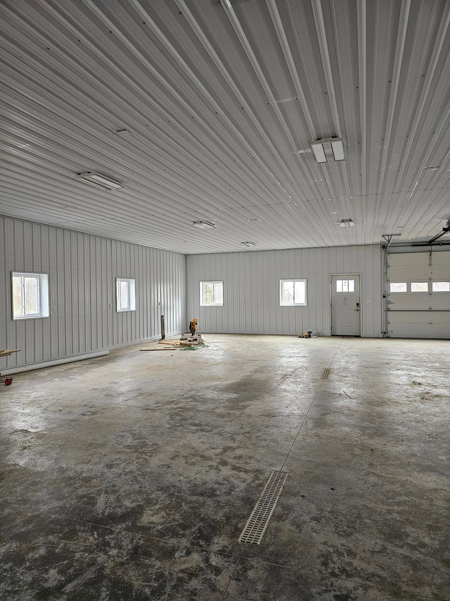 Empty industrial warehouse with corrugated metal walls and ceiling, concrete floor, windows, and a garage door.