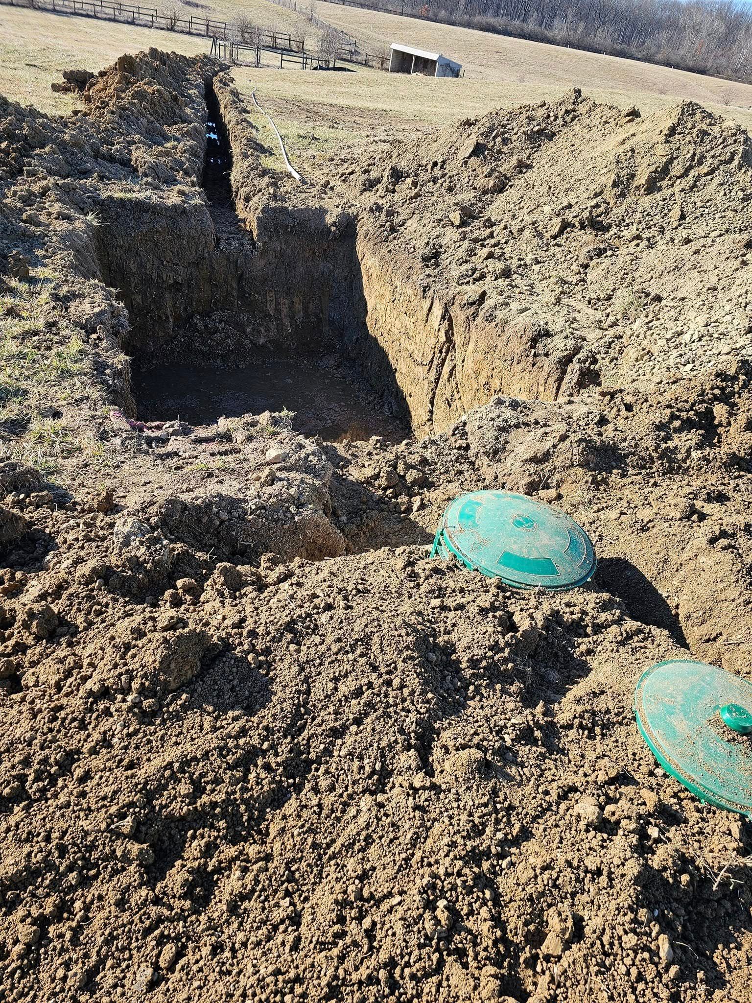 A hole in the ground revealing a septic tank, with two green lids visible, in a dirt field.