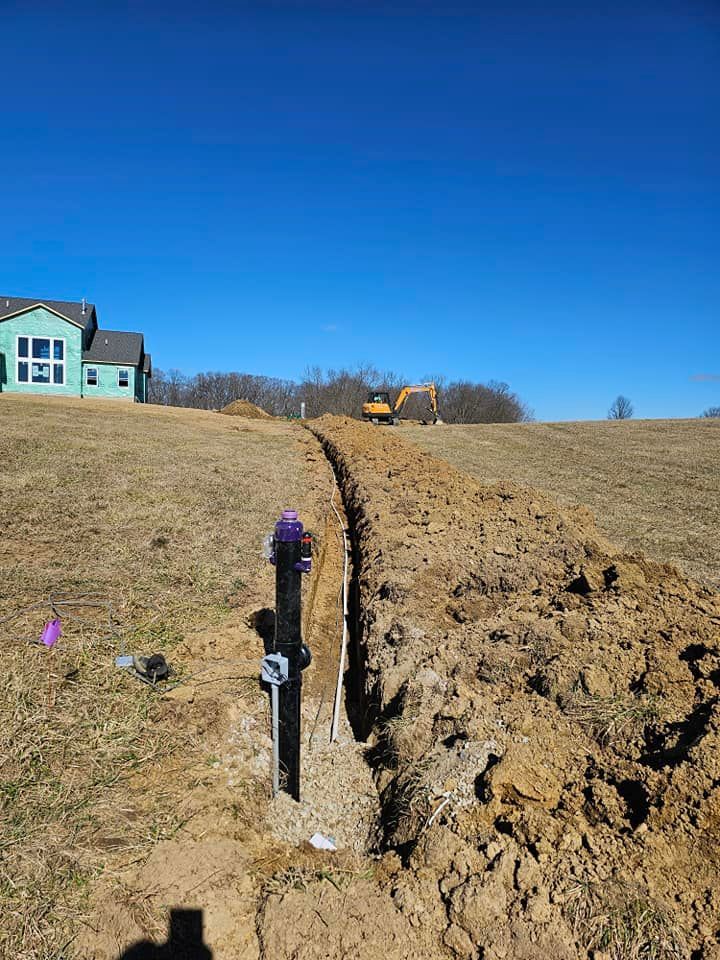 A trench dug in a field with a black pipe and construction in the background under a blue sky.