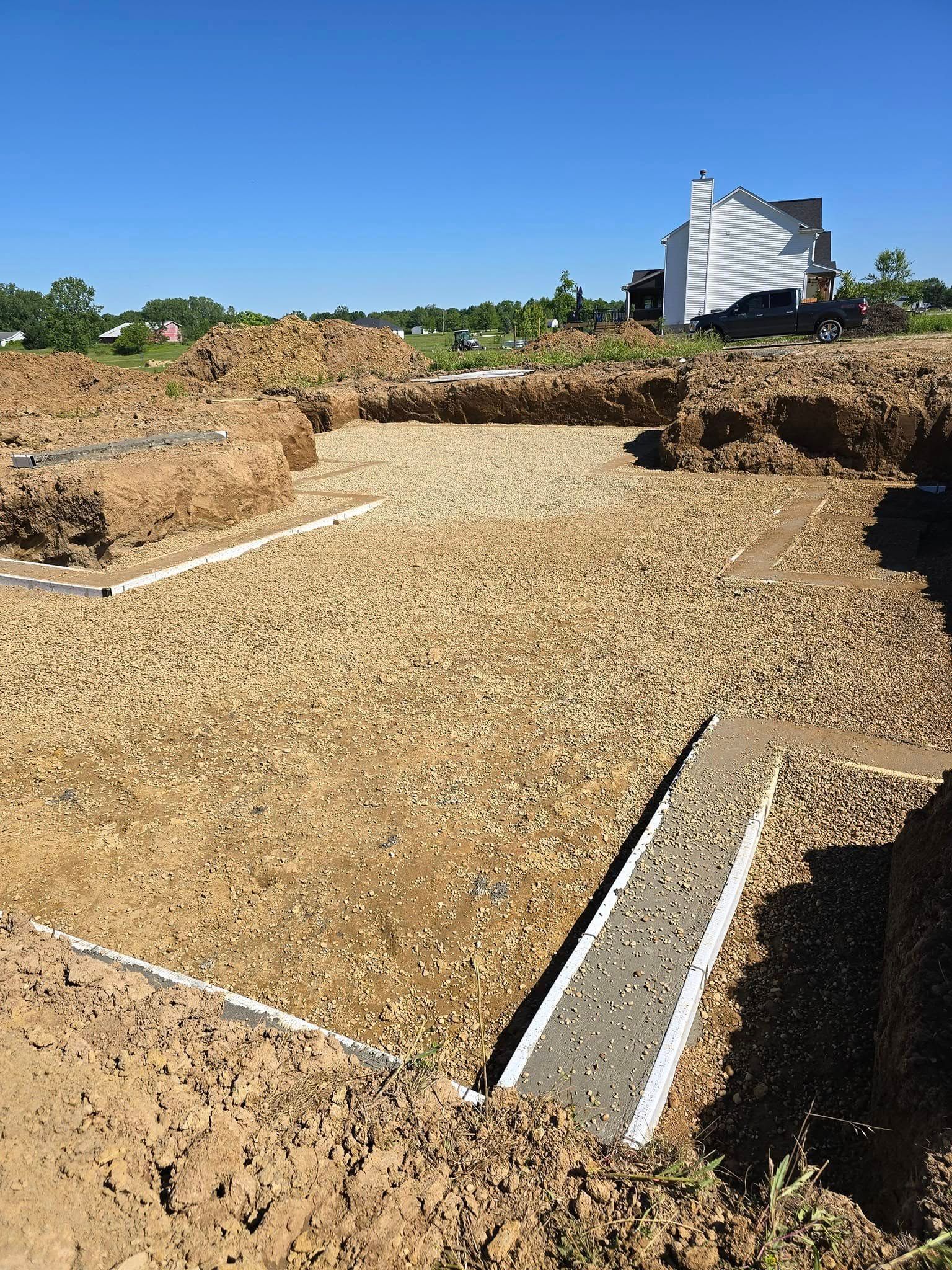 Excavated foundation site with gravel base, partially built concrete forms, and a distant white building under a blue sky.