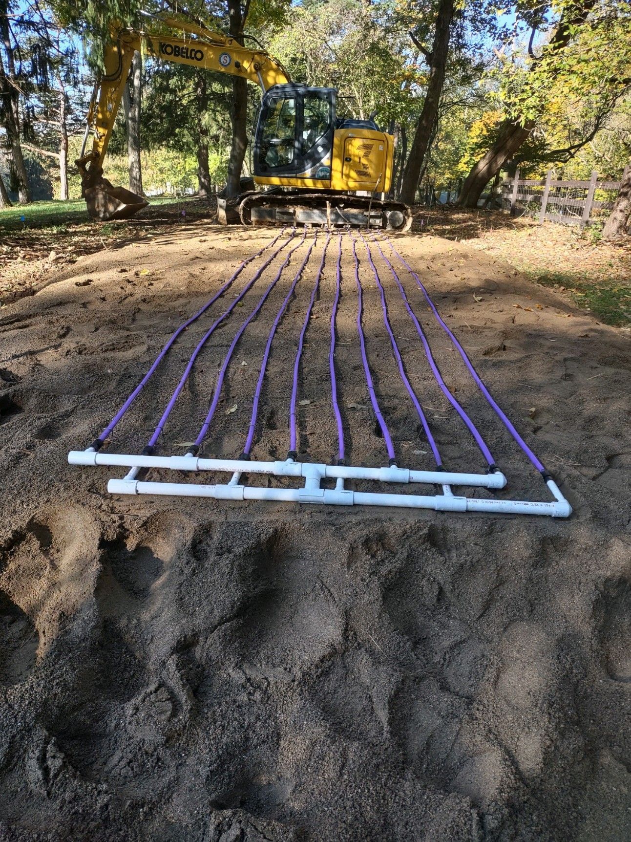 Purple pipes laid on soil, connected to white pipes. A yellow excavator is in the background, trees in the distance.