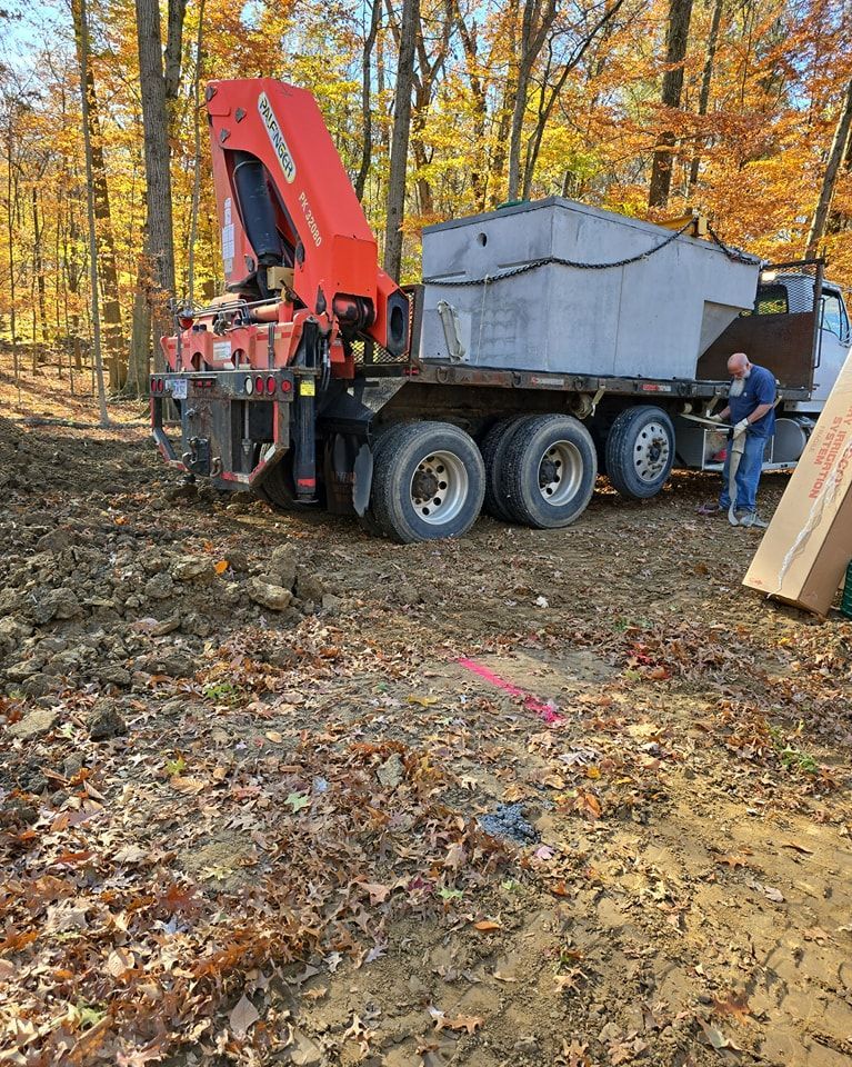 Crane truck unloading concrete structure in a wooded area; a worker is nearby.