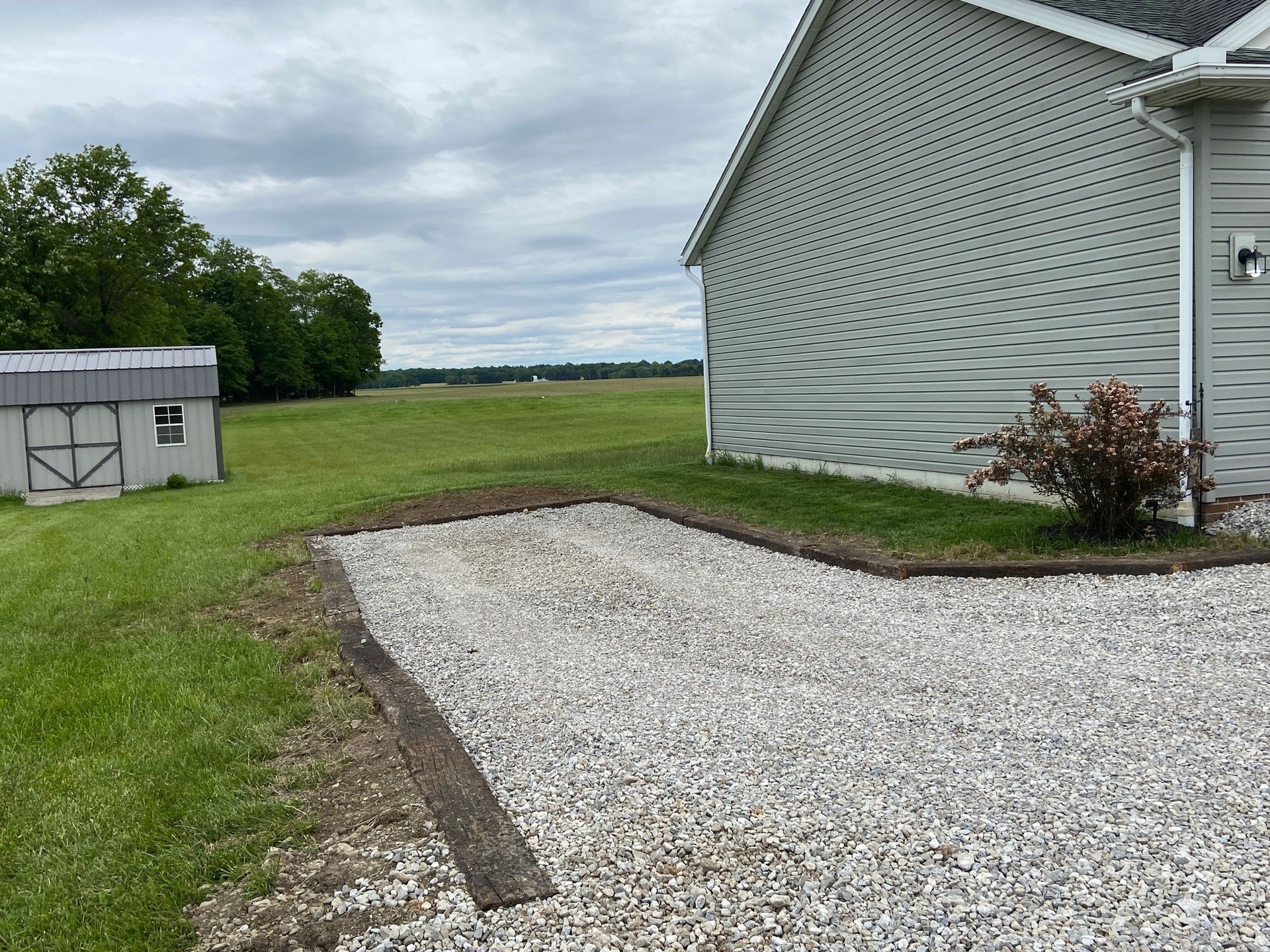 Gravel driveway edged with wood next to a house with a shed in a grassy field. Overcast day.