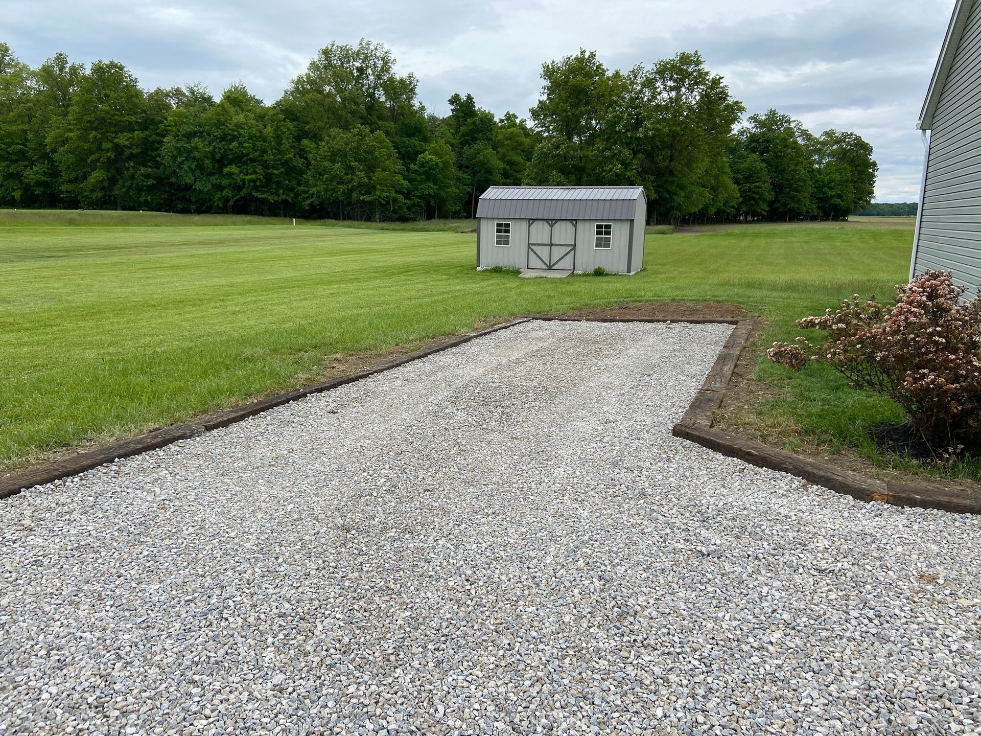 Gravel area with a shed in a grassy yard, bordered by wooden edging, with trees in the background.