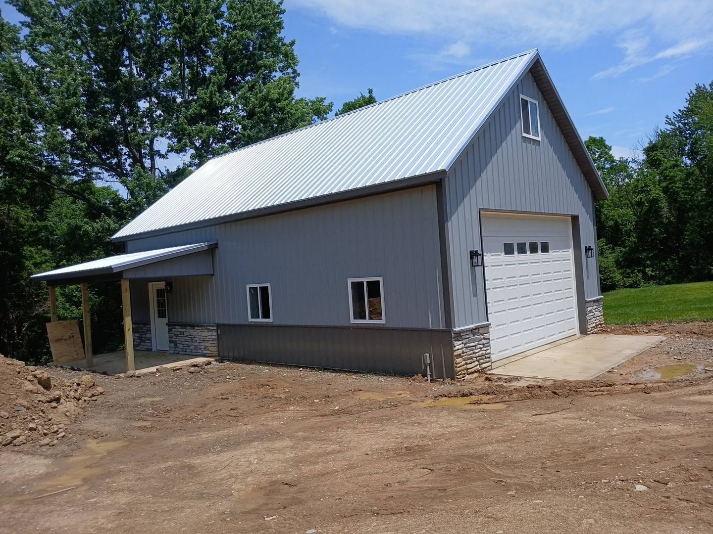 Gray barn with white garage door and metal roof, set in a clearing with trees.