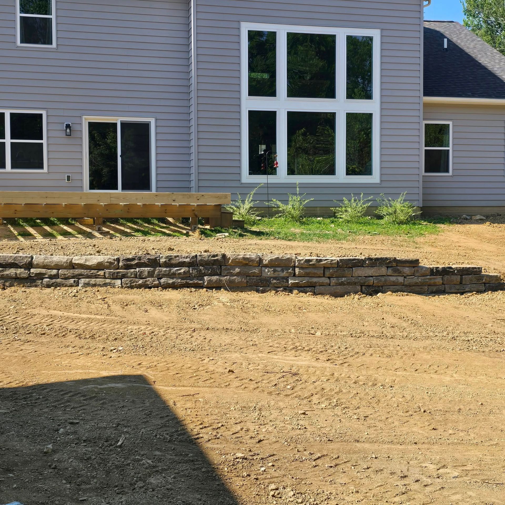 Back of a gray house with a retaining wall in front, on a graded dirt lot.