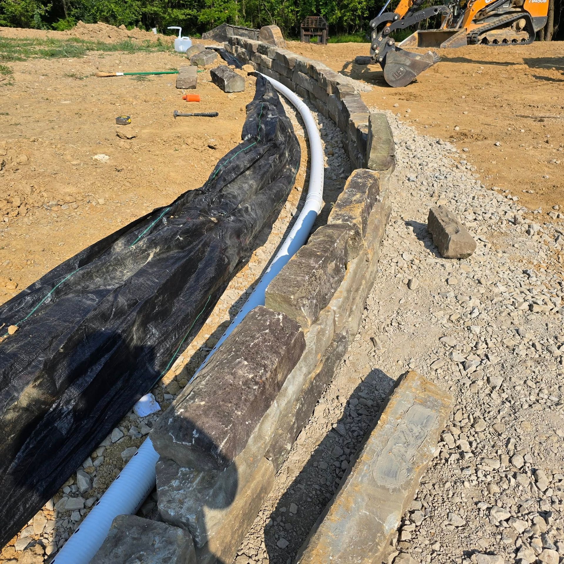 A trench with a white pipe, lined with stone and gravel, under construction outdoors.