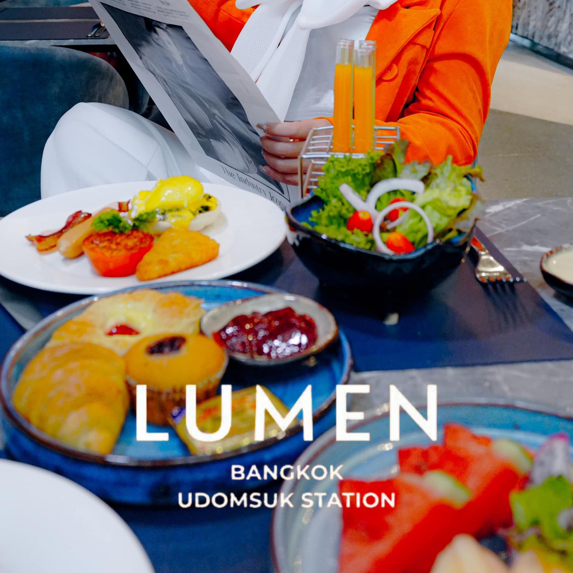 A woman is reading a newspaper while sitting at a table with plates of food and a bowl of salad in Lumen Hotel
