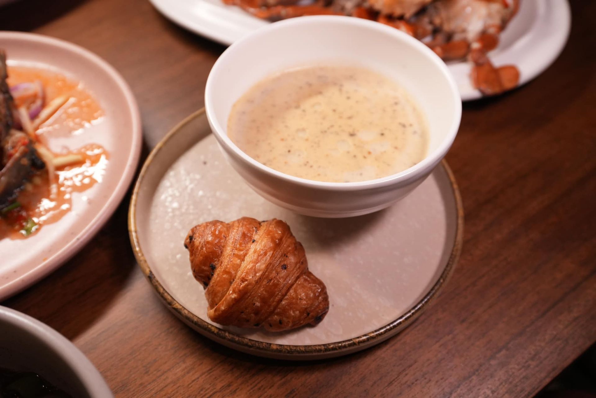 A bowl of soup and a croissant on a plate on a table.