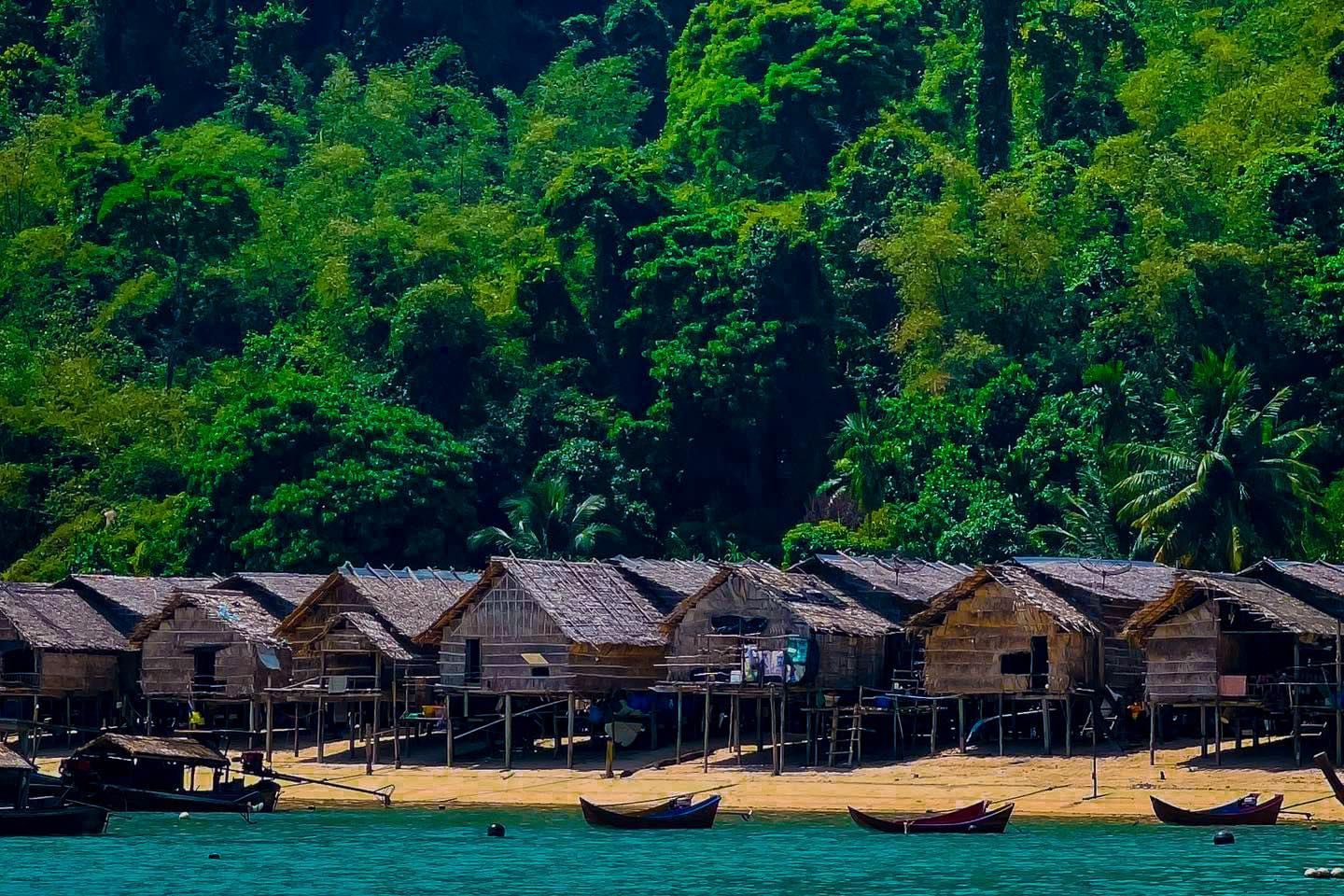 A row of thatched huts on stilts on a beach with boats in the water.