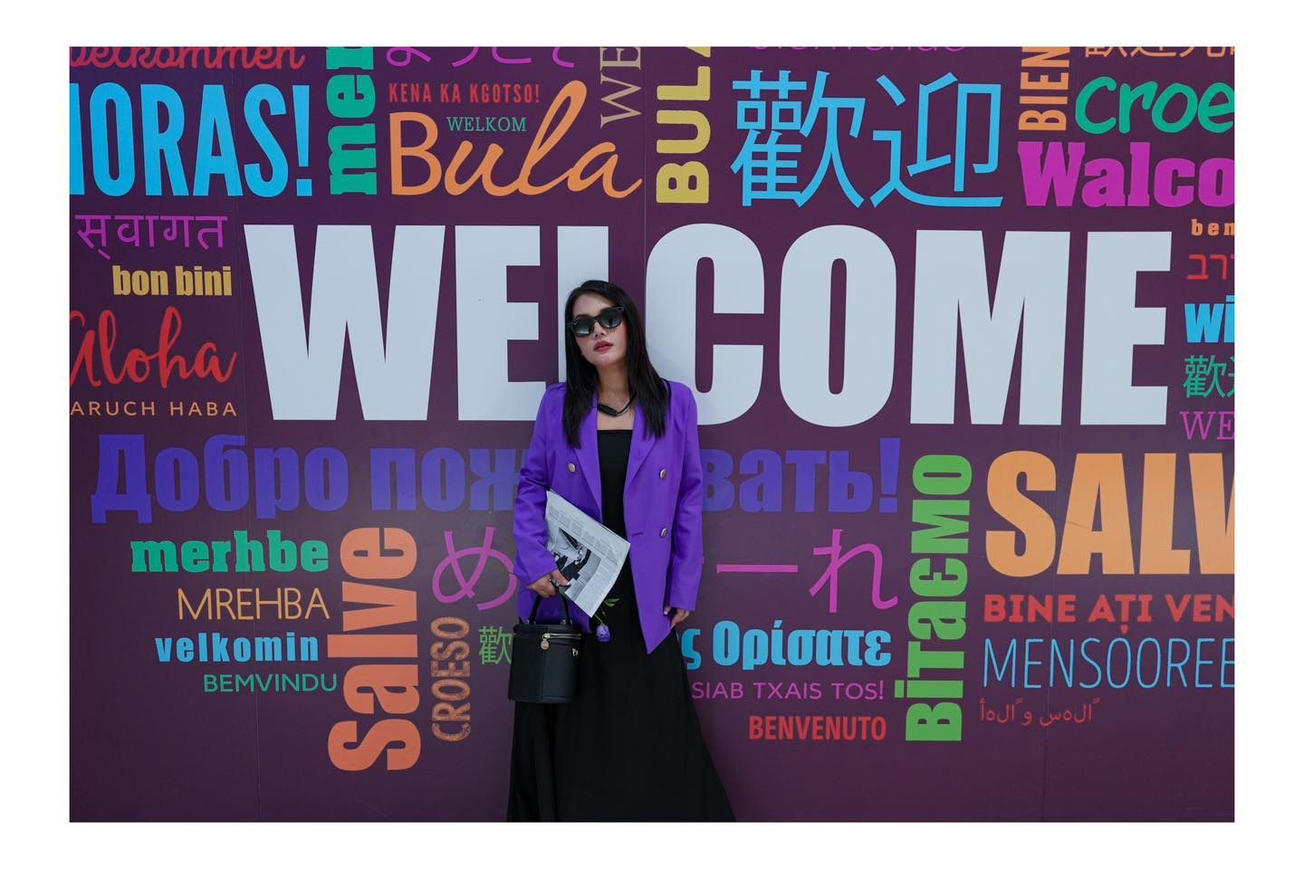 A woman is standing in front of a wall that says welcome in many different languages