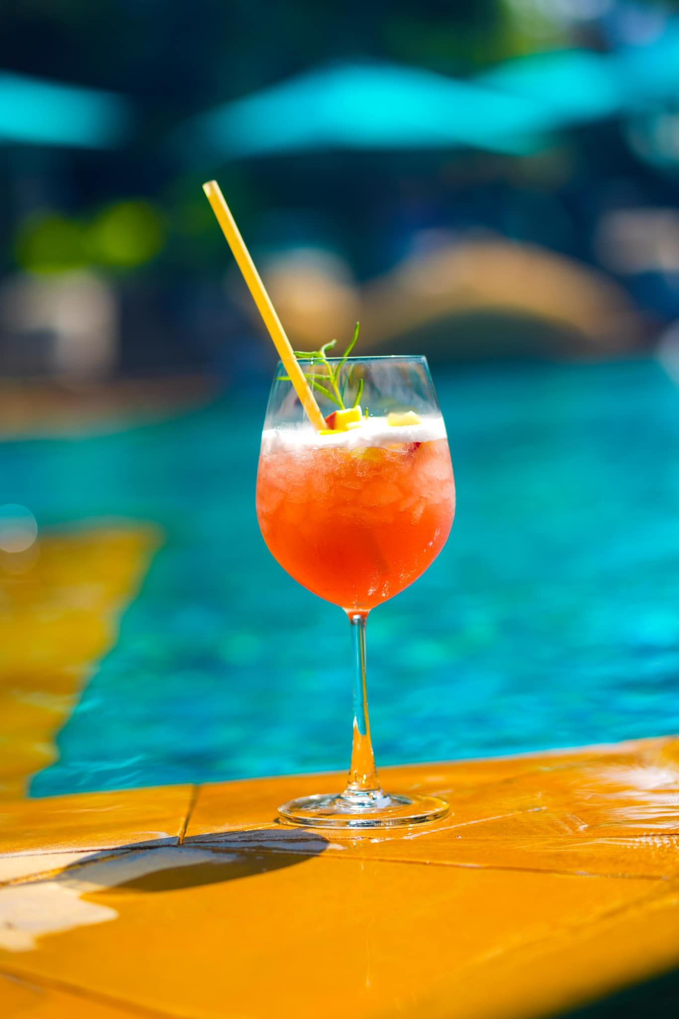 A red cocktail in a wine glass with a straw is sitting on the edge of a swimming pool.