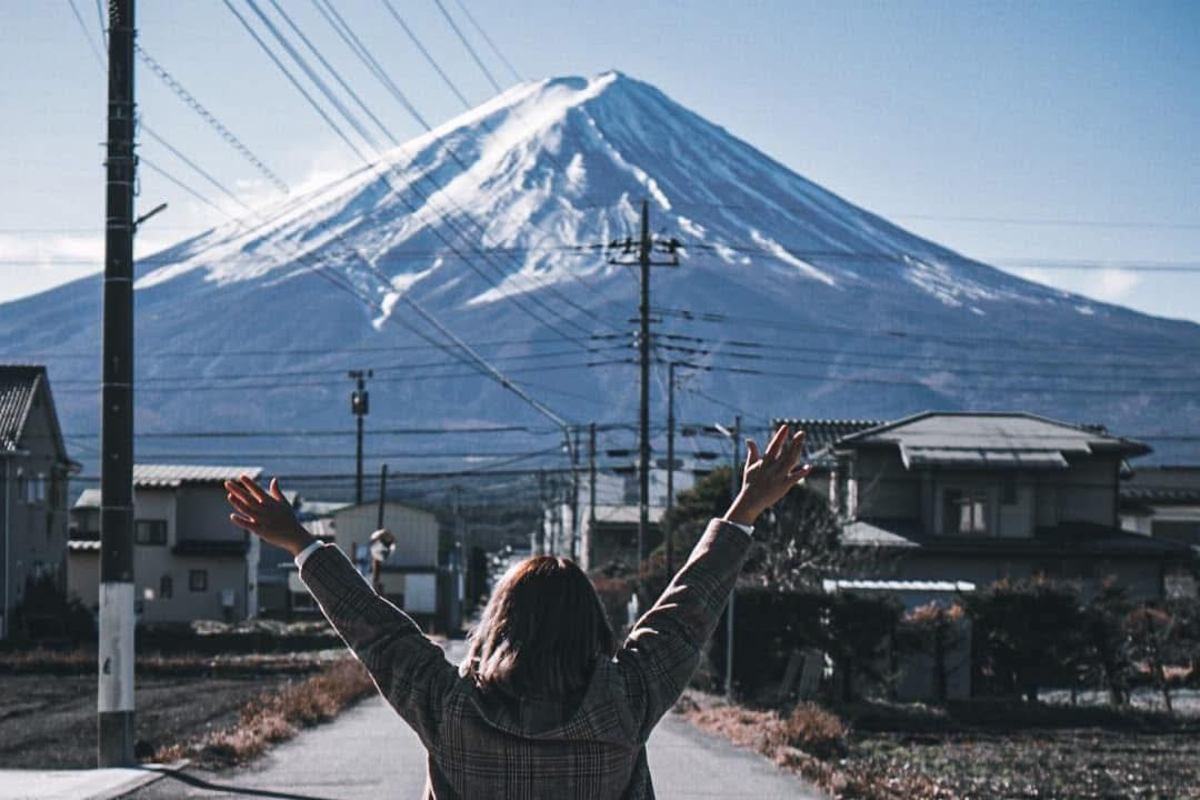 Woman with arms raised, facing Mt. Fuji, snowy peak, sunny day, small houses in the background.