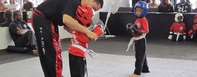 A martial arts instructor adjusts the headgear of a student in red gear, while another student watches nearby.