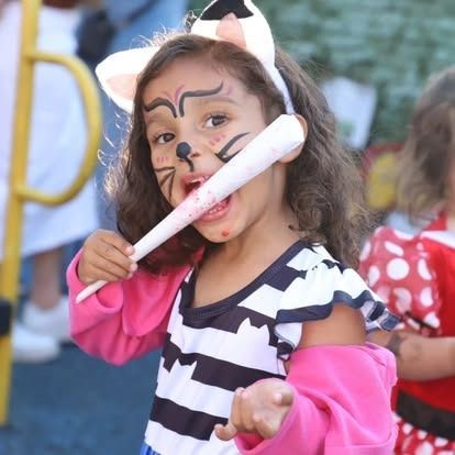 A child with cat face paint and ears holds a lollipop to their mouth, wearing a pink and black striped outfit.