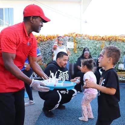A person in a red uniform and cap serves small snacks to two children outside, while a person in a skeleton suit watches.