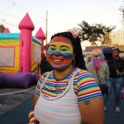 A smiling person with rainbow-painted face and matching striped shirt stands before a colorful inflatable bounce house.