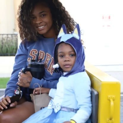 A smiling person sits next to a child wearing a blue dog-character hood and dress, sitting on a yellow structure.