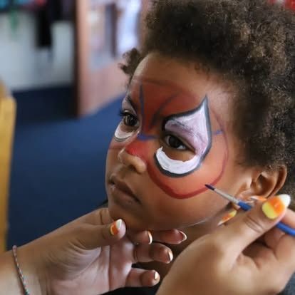 A person uses a thin brush to apply superhero-style face paint around a child's eyes.