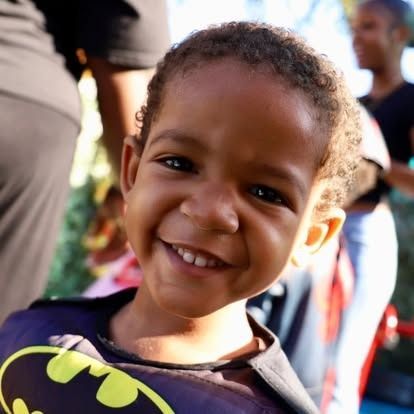 A close-up portrait of a smiling child wearing a dark purple Batman costume with a yellow logo.