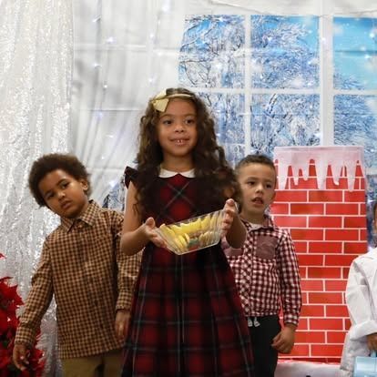 Three children stand in front of a holiday backdrop, with the central child holding a clear bowl of yellow snacks.