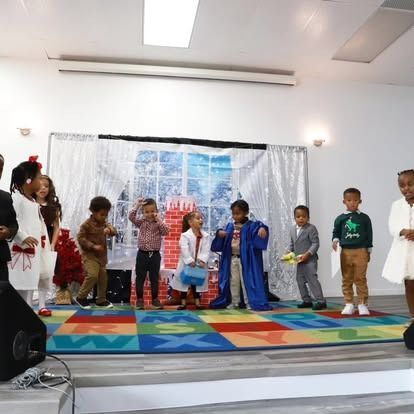 A group of children in costumes stand on a brightly colored alphabet rug in front of a holiday-themed backdrop.