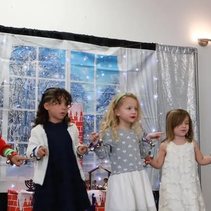 Three children stand in a line against a winter-themed backdrop, performing with jingle bells in their hands.