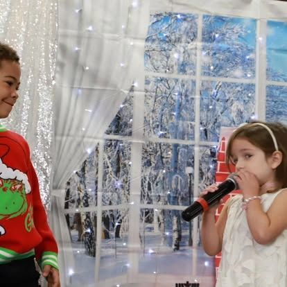 A child in a Grinch sweater looks at another child singing into a microphone in front of a wintry window backdrop.