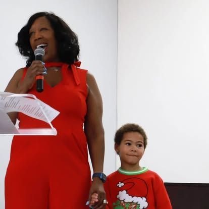 A woman in a red dress speaking into a microphone beside a child wearing a Grinch holiday sweater.